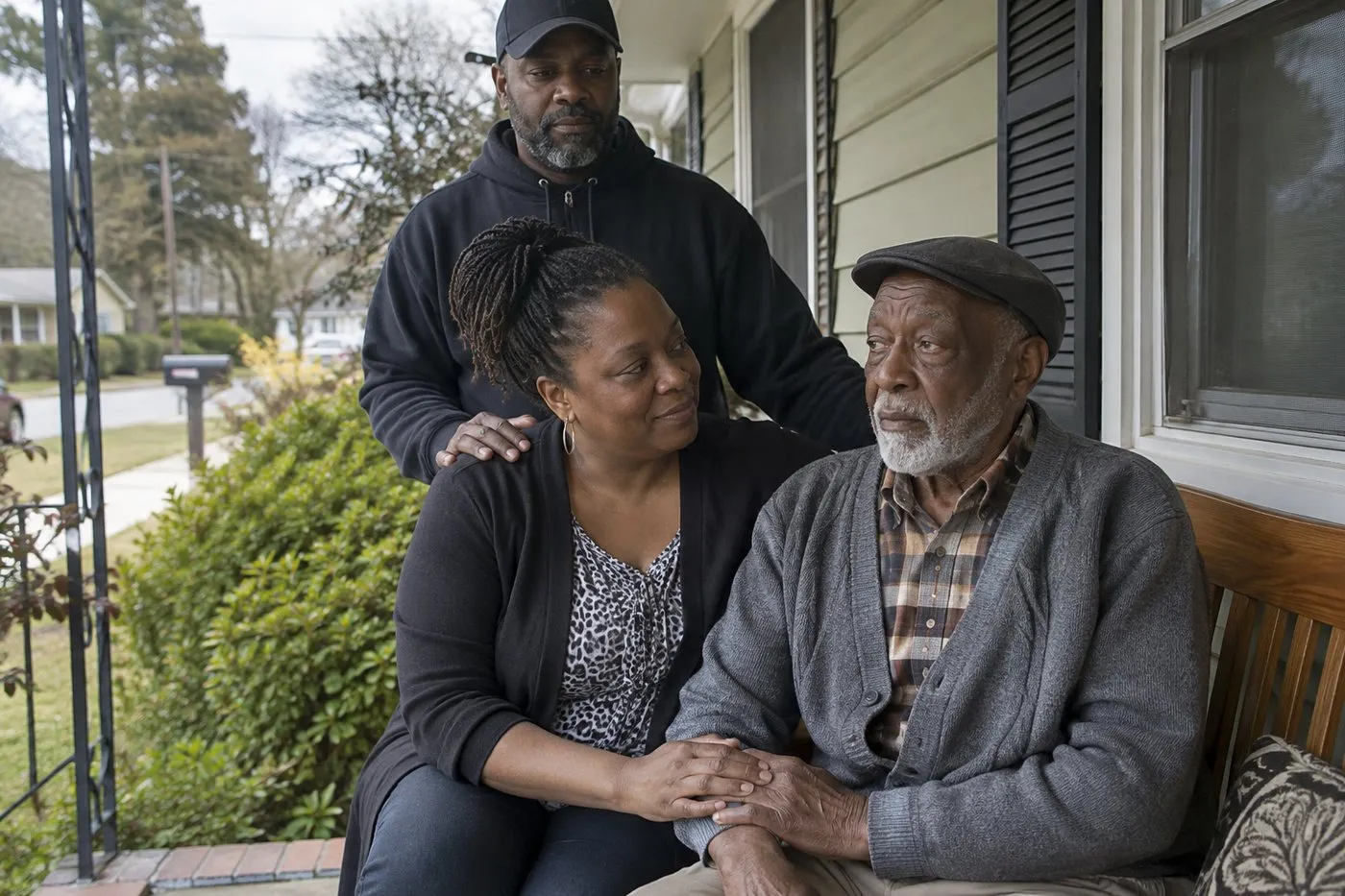 Adult daughter holding her elderly father's hand on a front porch bench while her husband stands behind them