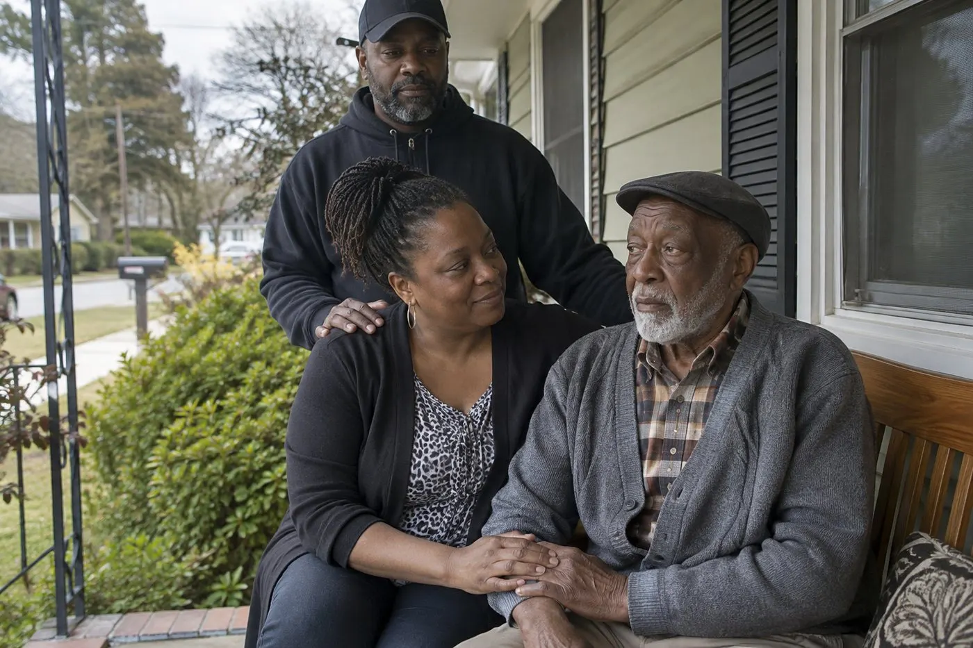 Adult daughter holding her elderly father's hand on a front porch bench while her husband stands behind them